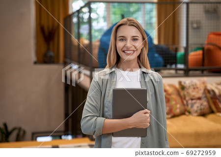 Portrait of a young cheerful caucasian woman, female office worker holding digital tablet and looking at camera while standing in the modern office Portrait of a young cheerful caucasian woman, female office worker holding digital tablet and looking at camera while standing in the modern office 69272959