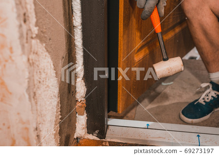The carpenter, after foaming and insulating the front door, knocks with a hammer for good adhesion of the foam to the wall and door. The carpenter, after foaming and insulating the front door, knocks with a hammer for good adhesion of the foam to the wall and door. 69273197