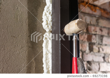 The carpenter, after foaming and insulating the front door, knocks with a hammer for good adhesion of the foam to the wall and door. The carpenter, after foaming and insulating the front door, knocks with a hammer for good adhesion of the foam to the wall and door. 69273198