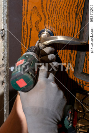 Close-up, the carpenter installs a custom lock in the front metal door, using a drill and hammer and other tools. Close-up, the carpenter installs a custom lock in the front metal door, using a drill and hammer and other tools. 69273205