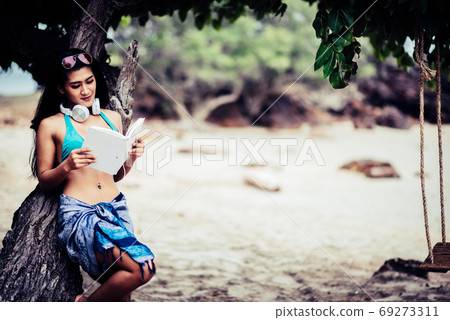 Young Woman Reading Book While Leaning On Tree At Beach Young Woman Reading Book While Leaning On Tree At Beach 69273311