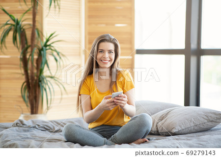 Smiling millennial girl with smartphone browsing web on bed at home Smiling millennial girl with smartphone browsing web on bed at home 69279143