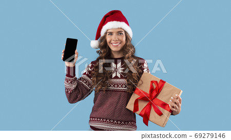 Smiling girl in Christmas outfit holding gift box and cellphone with blank screen over blue background, space for design Smiling girl in Christmas outfit holding gift box and cellphone with blank screen over blue background, space for design 69279146
