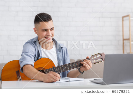 Smiling young man watching online lesson, playing guitar and makes notes Smiling young man watching online lesson, playing guitar and makes notes 69279231