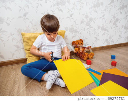 Toddler boy learns to cut colored paper with scissors. Kid sits on floor in kids room with toy blocks and teddy bear. Educational classes for children. Homeschooling. Toddler boy learns to cut colored paper with scissors. Kid sits on floor in kids room with toy blocks and teddy bear. Educational classes for children. Homeschooling. 69279781