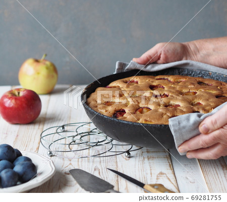 Hands of an elderly woman serve a frying pan with a ready made pie with apples and plums on a wooden white table. Homemade cakes. Hobby of a woman. Hands of an elderly woman serve a frying pan with a ready made pie with apples and plums on a wooden white table. Homemade cakes. Hobby of a woman. 69281755
