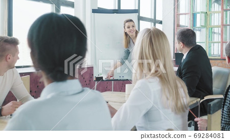 Business startup team holds a meeting at a large table. Girl manager gets up from a chair and walks over to a flip chart to start a presentation. Coworking. Office life Business startup team holds a meeting at a large table. Girl manager gets up from a chair and walks over to a flip chart to start a presentation. Coworking. Office life 69284081