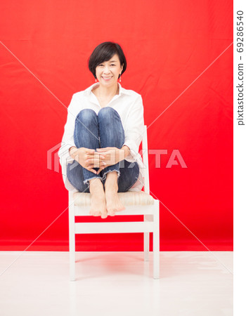 A middle-aged woman in a white shirt and jeans sitting on a chair red back A middle-aged woman in a white shirt and jeans sitting on a chair red back 69286540