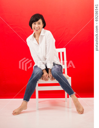 A middle-aged woman in a white shirt and jeans sitting on a chair red back A middle-aged woman in a white shirt and jeans sitting on a chair red back 69286541