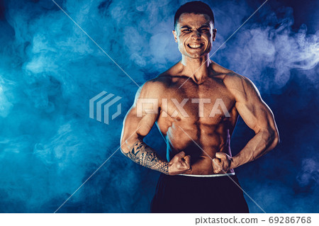Athletic man flexing muscles in studio on dark background with smoke. Strong bodybuilder with perfect abs 69286768