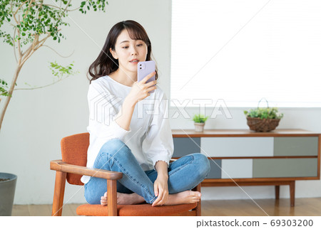 一個年輕的女人坐在客廳裡的椅子上,經營智能手機 一個年輕的女人坐在客廳裡的椅子上,經營智能手機 69303200