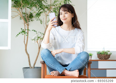 微笑著的年輕女子坐在客廳裡的椅子上和操作智能手機 69303209