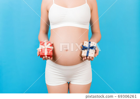 Cropped image of pregnant woman in white underwear holding two gift boxes at blue background. Is it a boy or a girl Pregnancy celebration. Copy space 69306261
