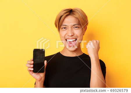 Close-up of happy rejoicing asian guy showing smartphone screen and saying yes, fist pump as triumphing, winning or achieve goal, yellow background Close-up of happy rejoicing asian guy showing smartphone screen and saying yes, fist pump as triumphing, winning or achieve goal, yellow background 69326579