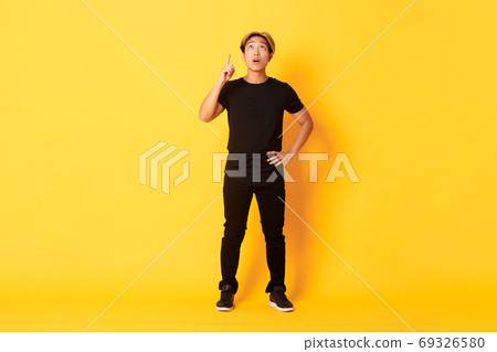 Full-length of thoughtful, intrigued attractive blond asian man raising finger, pointing up and looking amused, yellow background Full-length of thoughtful, intrigued attractive blond asian man raising finger, pointing up and looking amused, yellow background 69326580