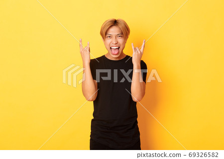 Portrait of joyful handsome asian blond guy having fun, showing rock-n-roll gesture and stick tongue carefree, standing yellow background Portrait of joyful handsome asian blond guy having fun, showing rock-n-roll gesture and stick tongue carefree, standing yellow background 69326582