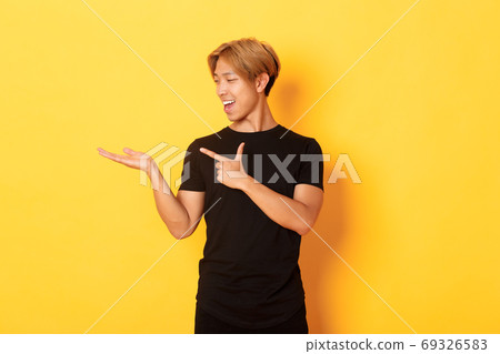 Portrait of handsome smiling asian guy holding something on hand and pointing finger at it, standing yellow background pleased Portrait of handsome smiling asian guy holding something on hand and pointing finger at it, standing yellow background pleased 69326583