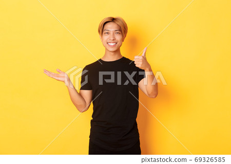 Satisfied and happy attractive korean guy smiling, showing thumbs-up in approval with rejoice, holding something on hand, standing yellow background Satisfied and happy attractive korean guy smiling, showing thumbs-up in approval with rejoice, holding something on hand, standing yellow background 69326585
