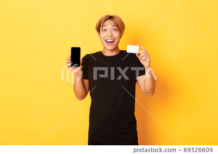 Portrait of excited happy asian man showing mobile phone screen and credit card with joyful smile, standing yellow background Portrait of excited happy asian man showing mobile phone screen and credit card with joyful smile, standing yellow background 69326609