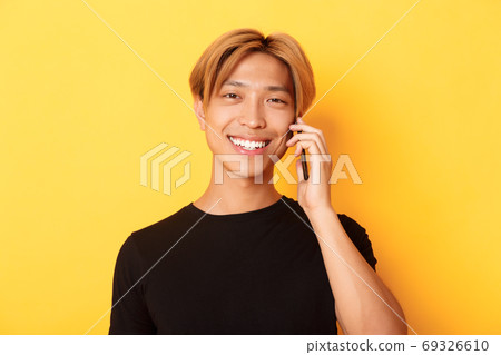Close-up of handsome stylish korean guy smiling and talking on mobile phone, standing over yellow background Close-up of handsome stylish korean guy smiling and talking on mobile phone, standing over yellow background 69326610