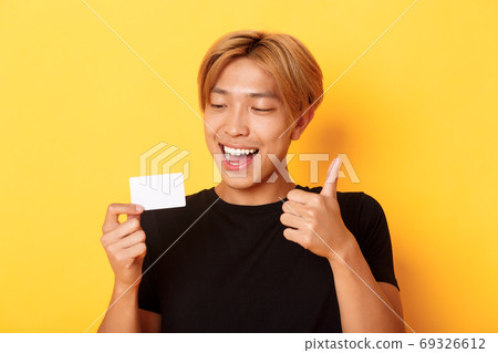 Close-up of happy and satisfied asian handsome guy, showing credit card and thumbs-up in approval, smiling amazed, standing yellow background 69326612