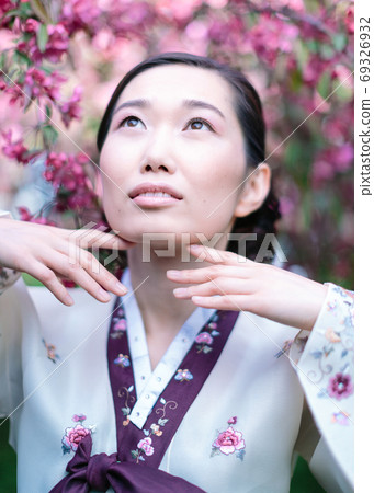 vertical portrait of an attractive Korean girl in a traditional costume with delicate make-up looks upwards, gently touching her fingers to her face on blossoming sakura background. vertical portrait of an attractive Korean girl in a traditional costume with delicate make-up looks upwards, gently touching her fingers to her face on blossoming sakura background. 69326932
