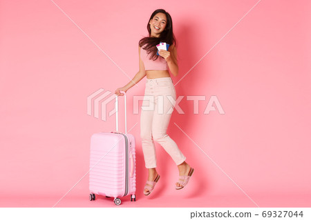 Travelling, holidays and vacation concept. Full-length of happy smiling asian girl, tourist with flight tickets and passport, jumping from excitement ready for summer paradise, pink background 69327044