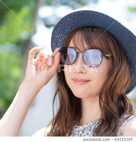 一個年輕的女人,戴著墨鏡,在初夏廣場的避暑勝地中微笑 一個年輕的女人,戴著墨鏡,在初夏廣場的避暑勝地中微笑 69333107