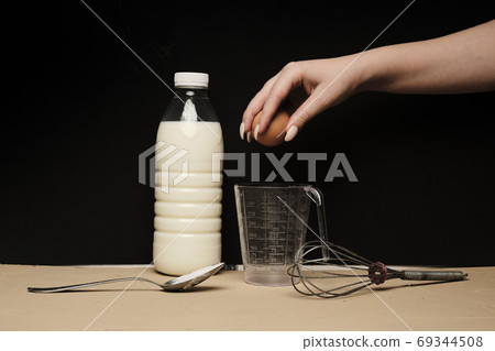 Female hands holding cracked egg over a white bowl, milk and kitchen tools on table 69344508
