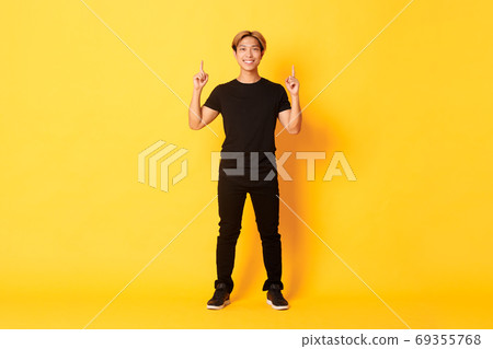 Full-length of confident handsome asian man with blond hair, smiling and pointing fingers up, showing logo, yellow background Full-length of confident handsome asian man with blond hair, smiling and pointing fingers up, showing logo, yellow background 69355768