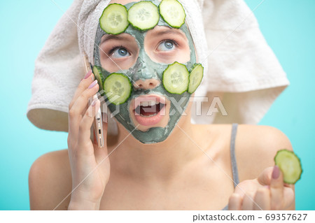 Woman with towel on head and face mask enriching with cucumbers via smartphone Woman with towel on head and face mask enriching with cucumbers via smartphone 69357627