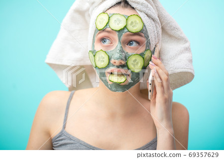 Woman with towel on head and face mask enriching with cucumbers via smartphone 69357629