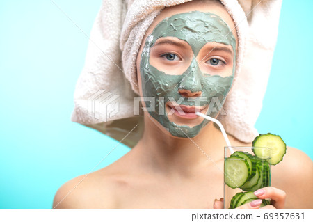 woman with cosmetic clay mask drinking cucucmber water isolated at studio blue woman with cosmetic clay mask drinking cucucmber water isolated at studio blue 69357631