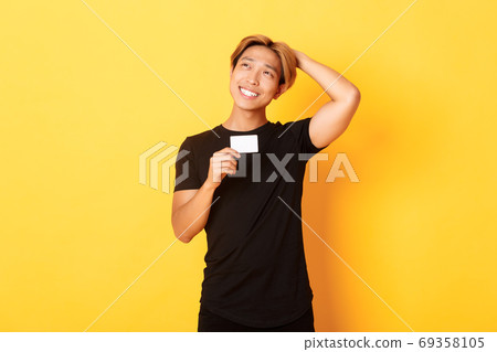 Thoughtful smiling asian guy thinking while showing credit card, looking upper left corner dreamy, yellow background 69358105