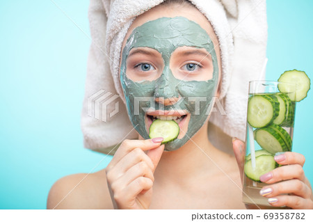 Positive smiling girl with clay cosmetic mask and organic cucumber slices 69358782