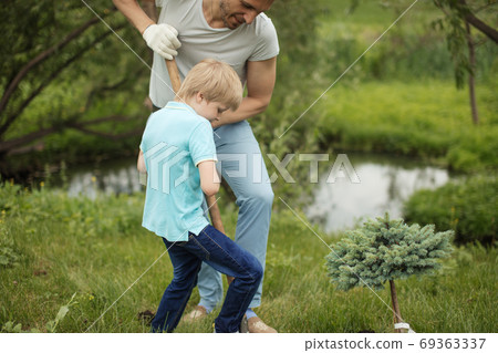Smiling adult father and his son planting a tree outdoors in park. Smiling adult father and his son planting a tree outdoors in park. 69363337