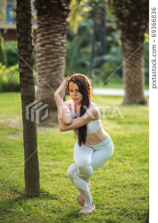 Woman in white sports outfit performing Yoga Eagle pose outdoor, in park meadow. 69366836