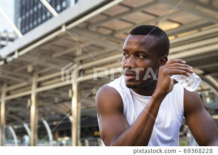 portrait of relaxing young african american sportsman sitting and drinking water from bottle after running exercise in city in morning with building background. sports exercise and city lifestyle. portrait of relaxing young african american sportsman sitting and drinking water from bottle after running exercise in city in morning with building background. sports exercise and city lifestyle. 69368228
