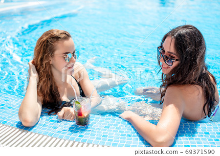 Two attractive brunette and blonde girls with long hair posing near pool on the sun. 69371590