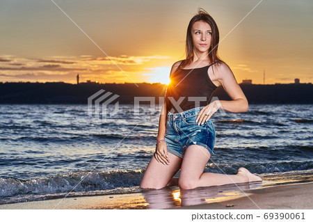 A young brunette girl in a black T-shirt and denim shorts kneels on the sandy shore  69390061