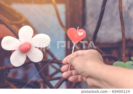 A red heart-shaped candle on woman's hand and an artificial flower made of white and red candles with bluerd image of rusty steel rod in the background. A red heart-shaped candle on woman's hand and an artificial flower made of white and red candles with bluerd image of rusty steel rod in the background. 69400124
