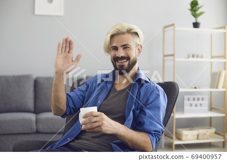 Smiling man sitting at home and greeting somebody with hand online looking at web camera Smiling man sitting at home and greeting somebody with hand online looking at web camera 69400957