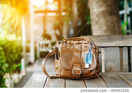 Mini portable alcohol gel bottle to kill Corona Virus(Covid-19) hang on a brown leather shoulder bag on table in garden. New normal lifestyle. Health care concept. Selective focus on alcohol gel 69401058