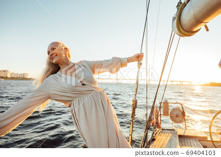 Cheerful senior woman in long dress enjoying vacation on private sailboat 69404103