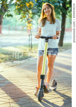 Smiling woman rides electric scooter or e-scooter in city park at sunset. Female using electric transport in urban park 69404634