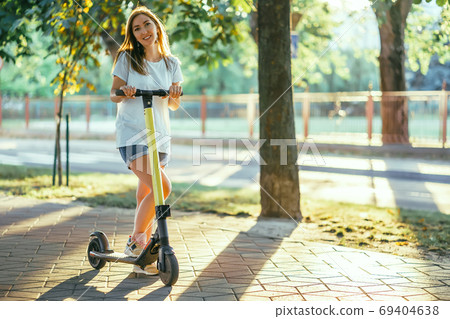 Smiling woman rides electric scooter or e-scooter in city park at sunset. Female using electric transport in urban park Smiling woman rides electric scooter or e-scooter in city park at sunset. Female using electric transport in urban park 69404638