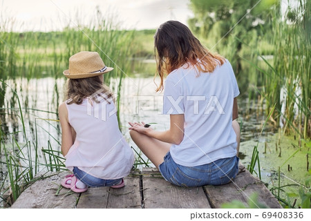 Children resting near the water on summer day, playing with water snails, back view Children resting near the water on summer day, playing with water snails, back view 69408336