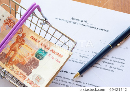 There is a bundle of money in a grocery basket and a ballpoint pen on the land purchase agreement 69421542
