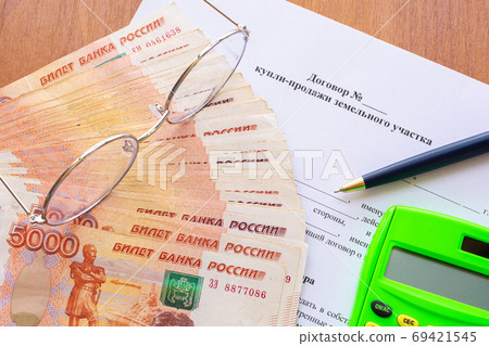On the table is a contract for the sale of a land plot, a bundle of money, a calculator, glasses and a pen 69421545