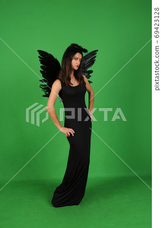 Beautiful young brunette woman in black angel costume with wings over green background. Halloween party outfit 69423128